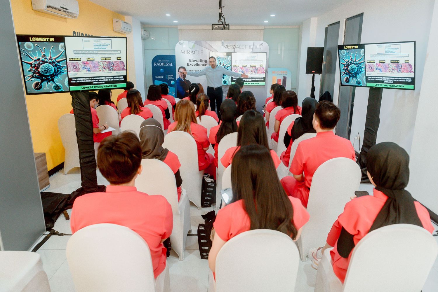 Students in red and white uniforms watch multiple screens during a classroom training session.