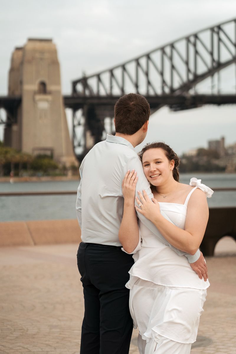 A couple shares a romantic moment near the iconic Sydney Harbour Bridge on an overcast day.