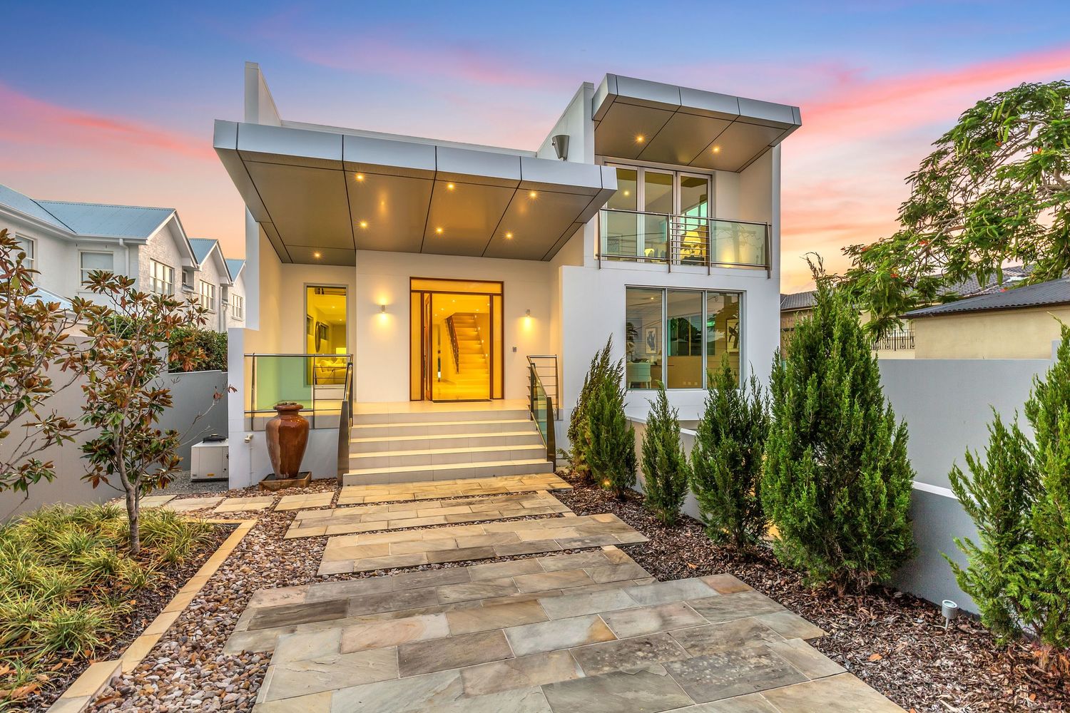 Stunning contemporary two-story home with illuminated entrance, manicured gardens, and warm sunset sky in Australia.