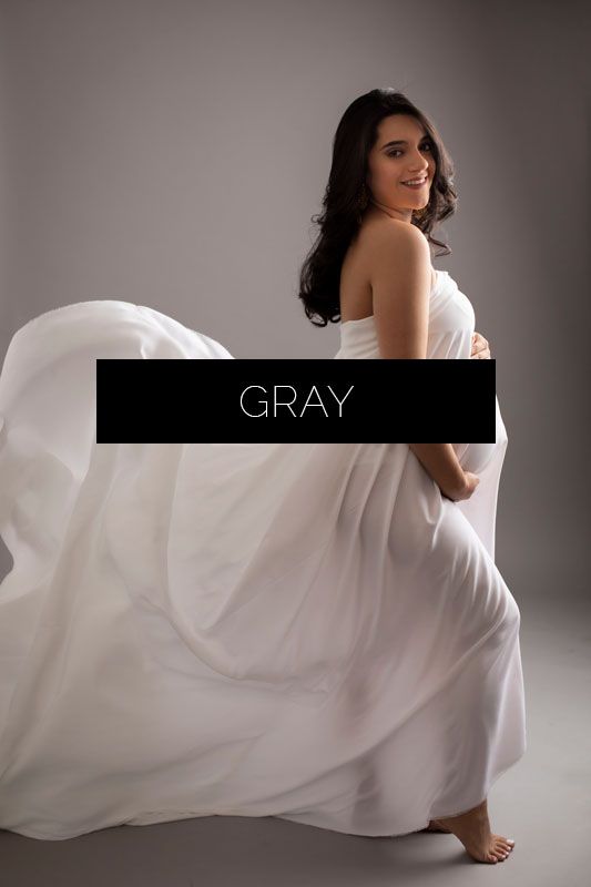 Artistic maternity photo featuring flowing white fabric against a gray studio backdrop with dramatic lighting.