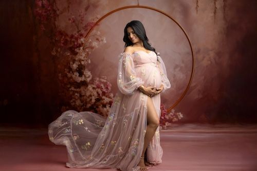 Pregnant woman in sheer pink floral maternity gown posing elegantly in front of a circular gold hoop with floral backdrop.