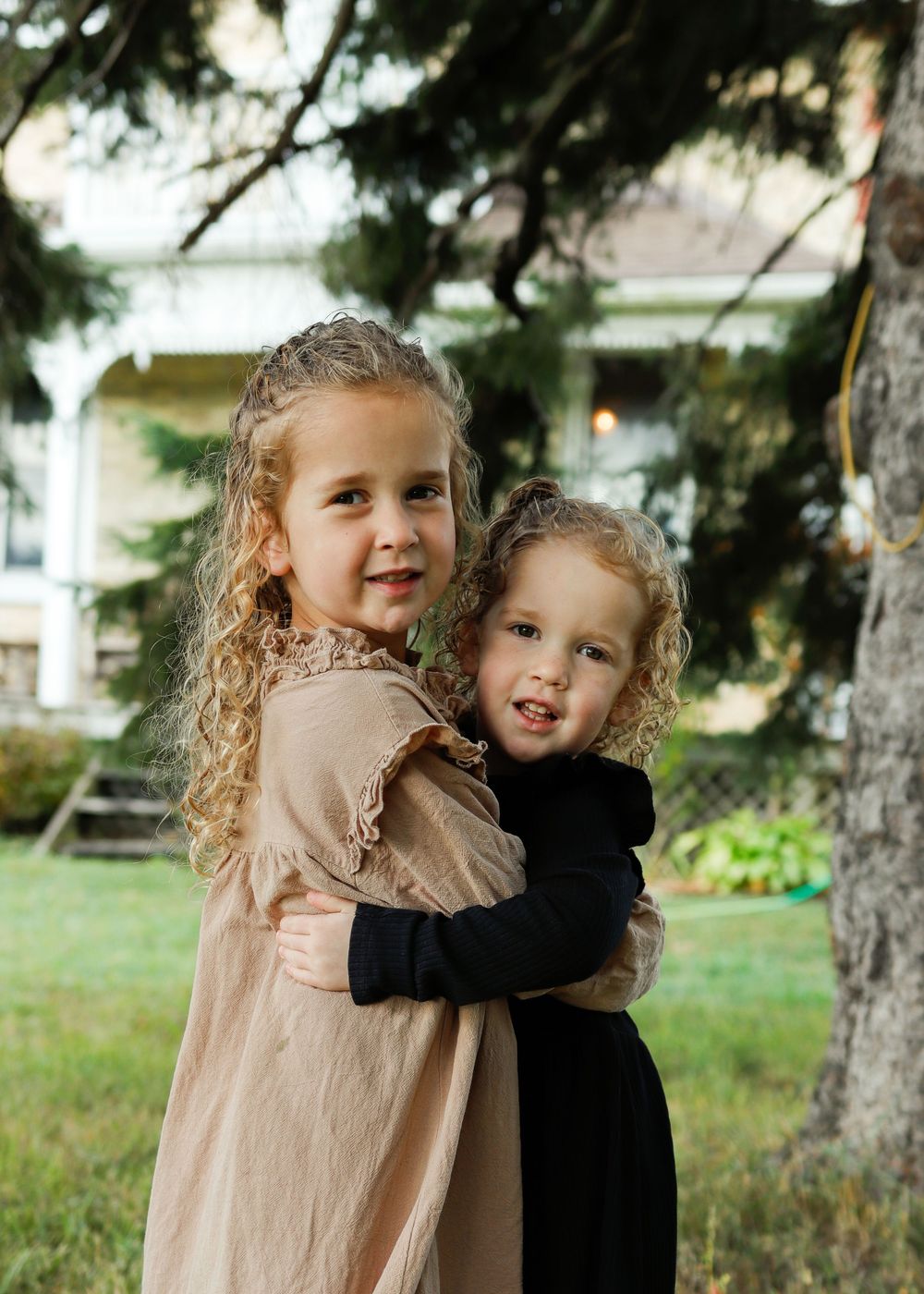 Two children with curly hair embrace outdoors, wearing contrasting beige and black dresses in front of a house.