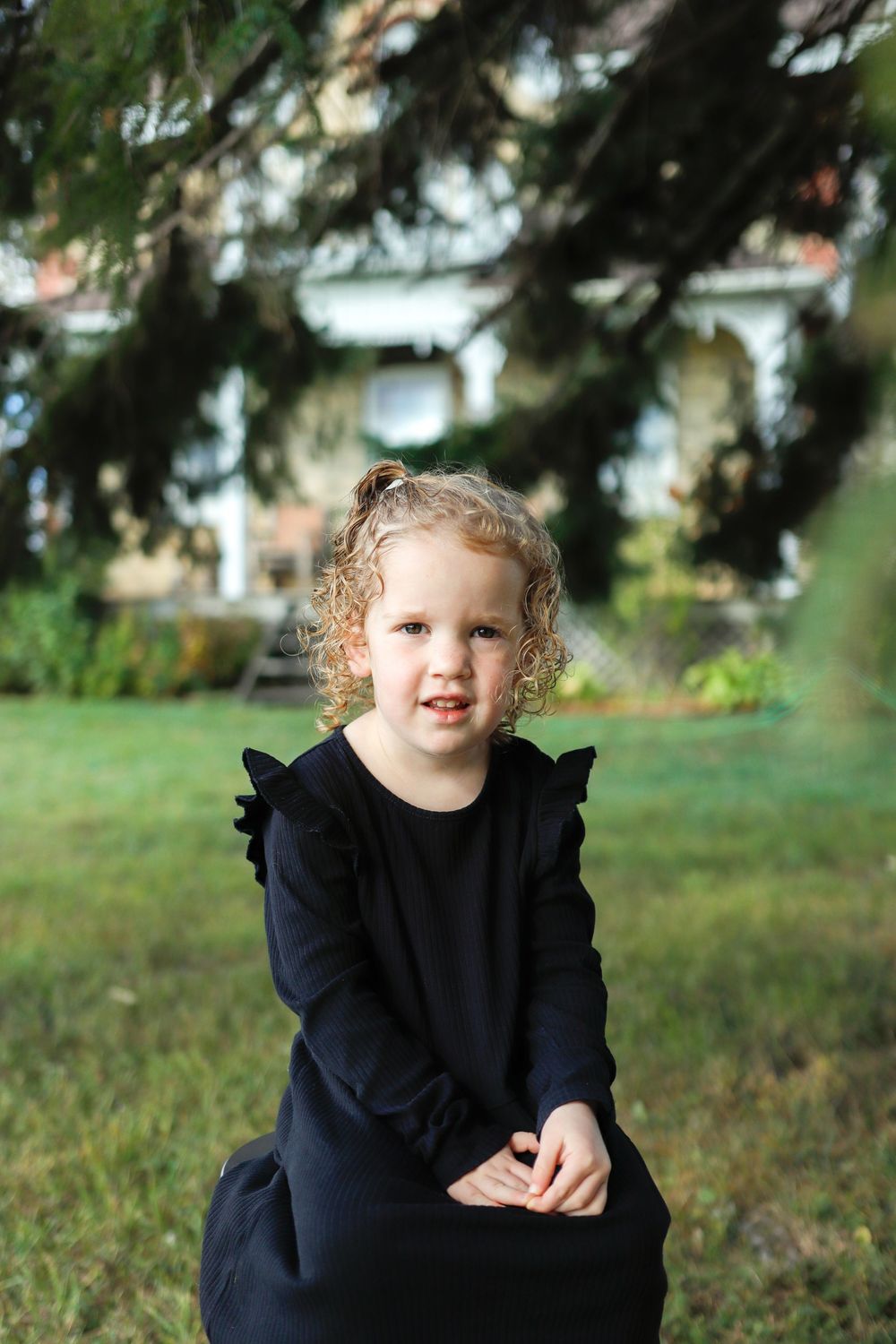A young child with curly hair wearing a black ruffled dress sits and plays outdoors on grass in a series of candid shots.