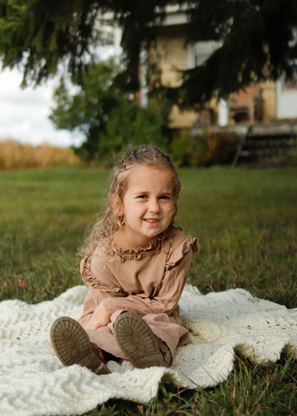 A child in a beige sweater lies on a white blanket in the grass during an outdoor photoshoot.