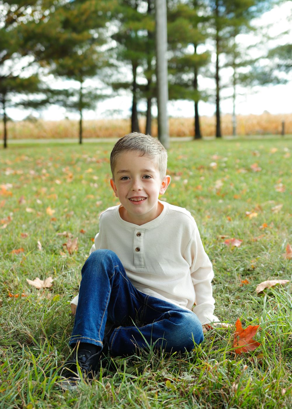 A child sits on autumn grass wearing a cream henley sweater and jeans with pine trees in the background.