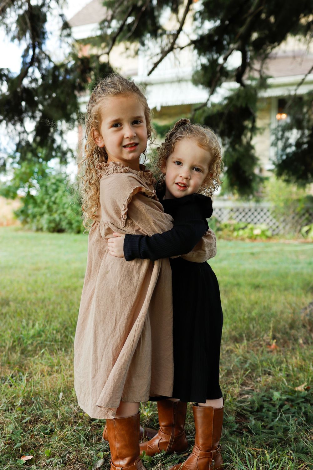 Two young children in brown boots embrace while playing outdoors on a grassy lawn.