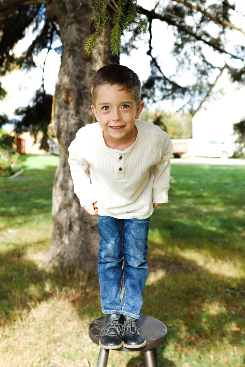 A young child in a white henley shirt and jeans stands on a small platform under a tree in a sunny outdoor setting.