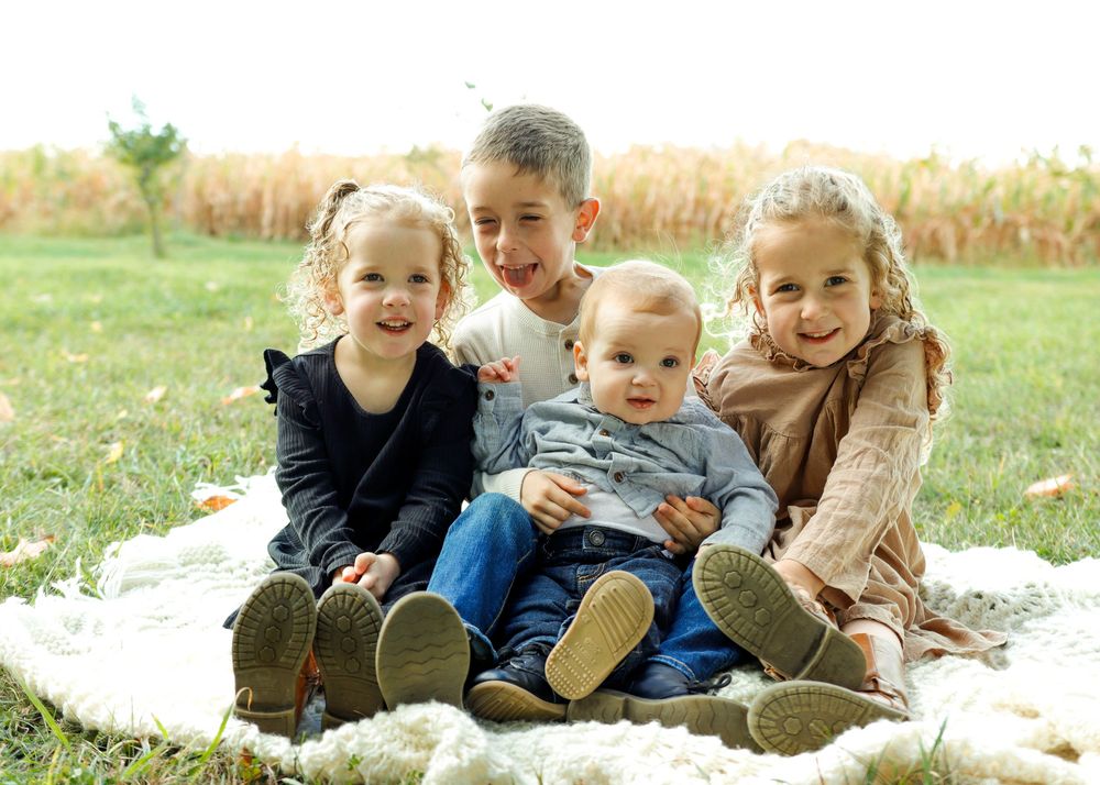 Children sitting together on a white blanket in a grassy field with corn stalks visible in the background.
