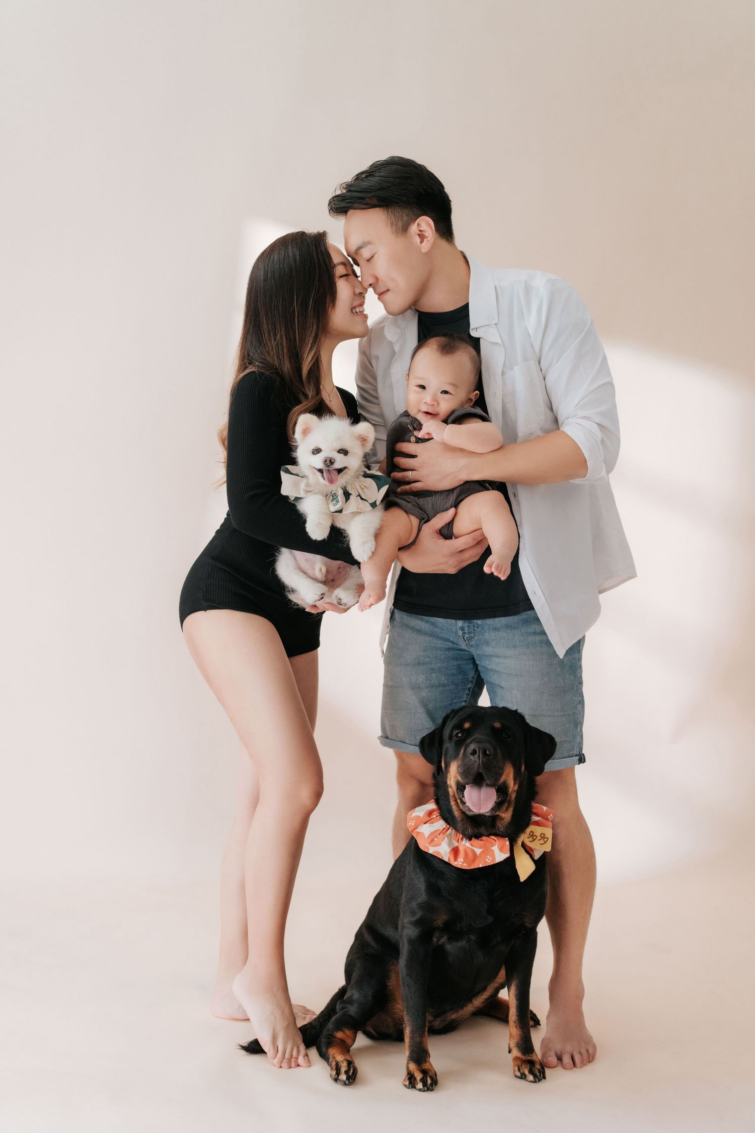 Young Asian couple sharing a kiss while holding a baby and small white dog, with a Rottweiler sitting in front.一對年輕的亞洲夫婦抱著嬰兒和一隻白色小狗，深情地擁吻