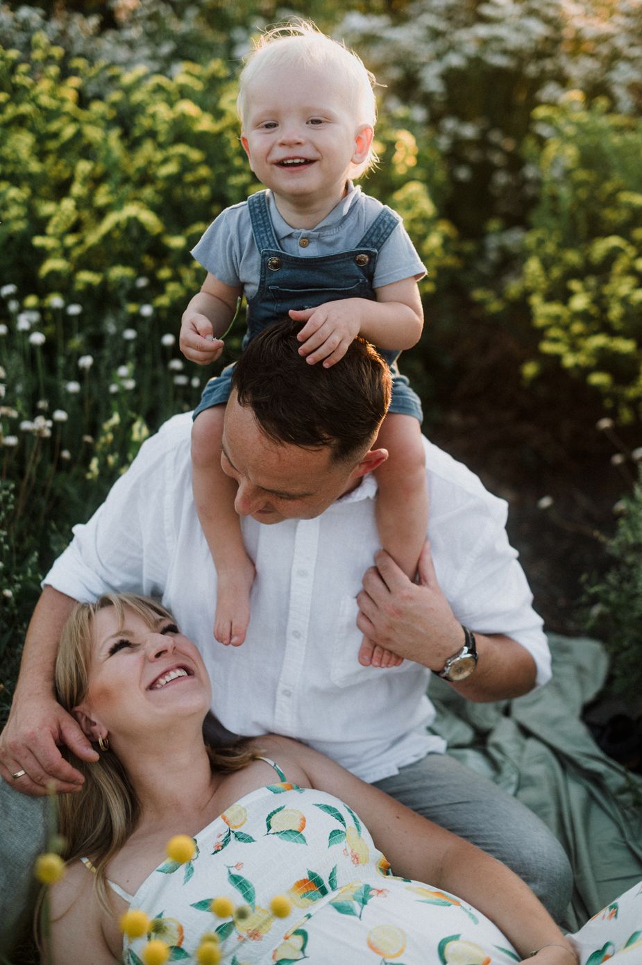 A joyful family moment captured outdoors with sunlight filtering through trees and yellow flowers in the background.