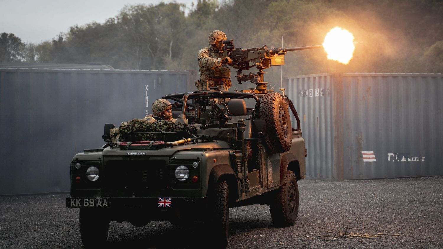 Military vehicle convoy moves through a dusty road at sunset with bright lights illuminating the path ahead.