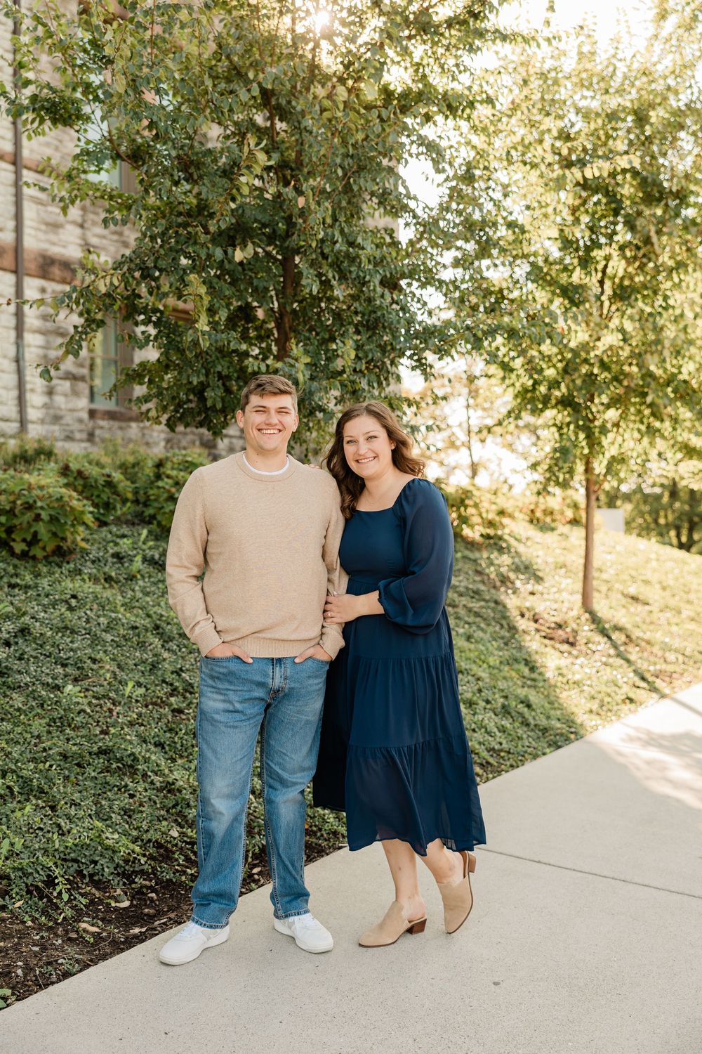 A couple poses together outdoors on a paved pathway surrounded by greenery.