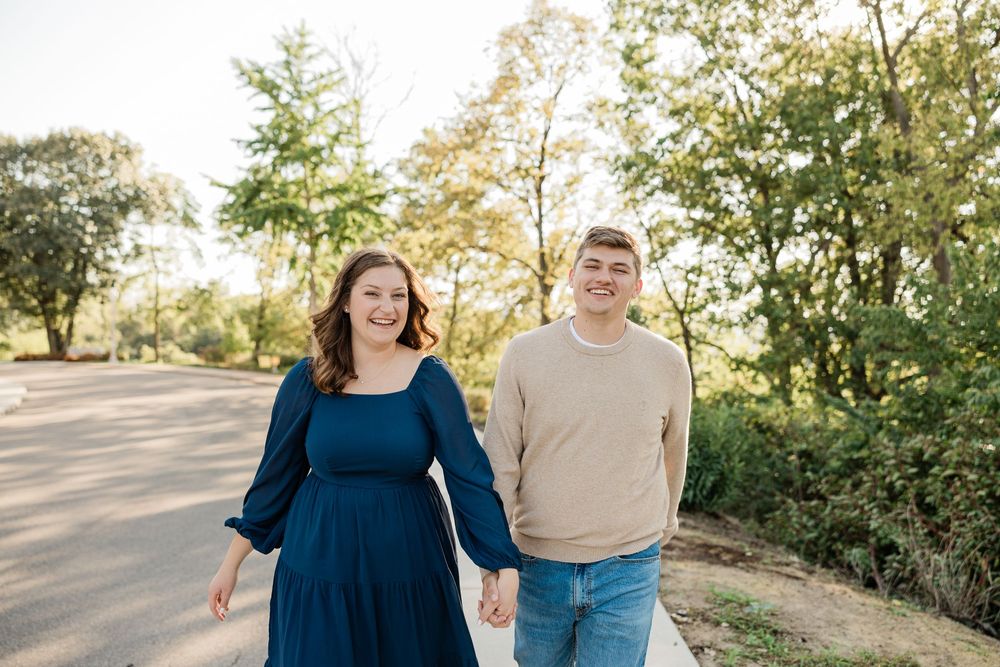 A couple walks hand in hand along a tree-lined path on a sunny day.