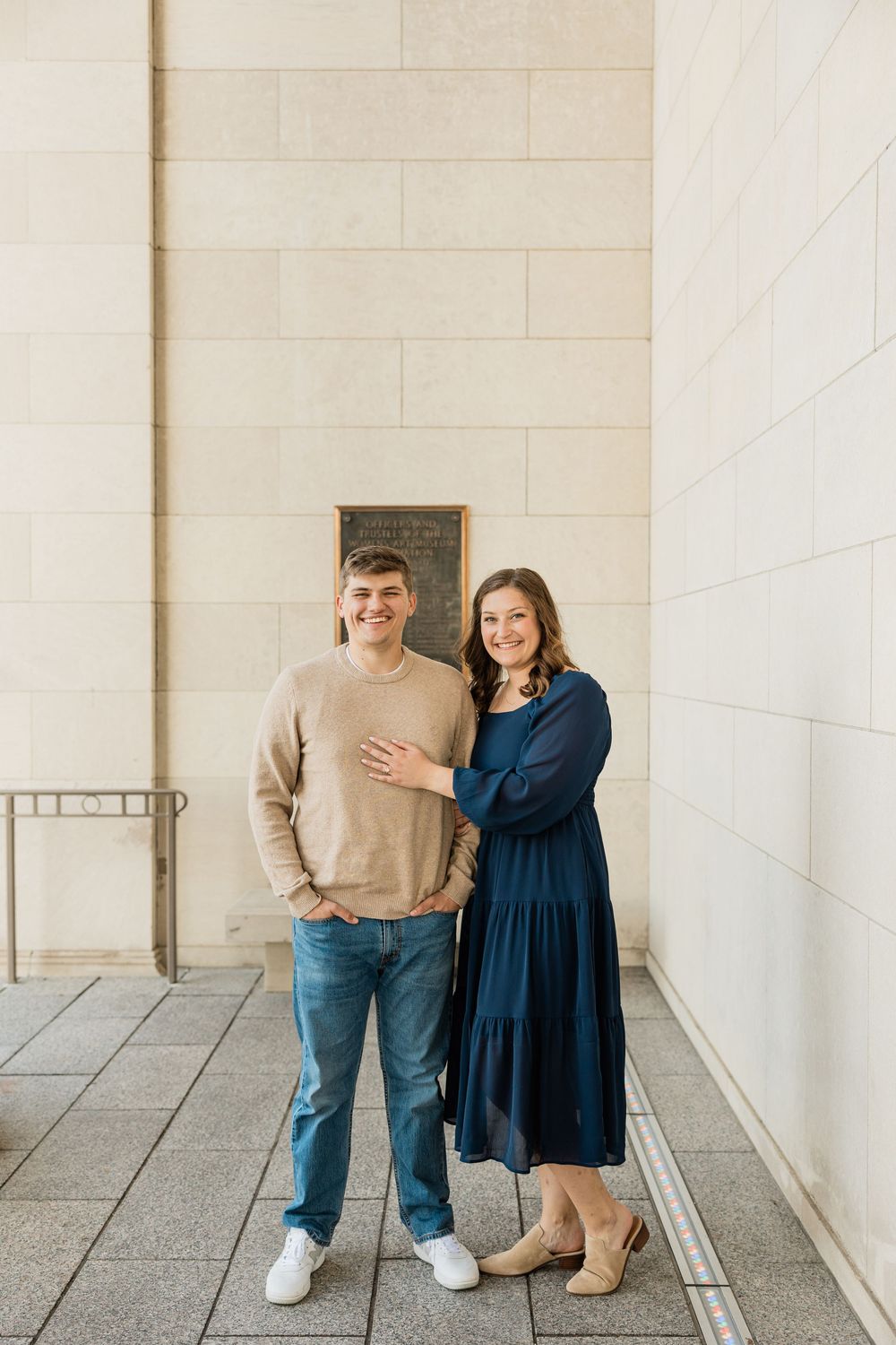 A couple stands together against a white wall in a modern architectural setting.