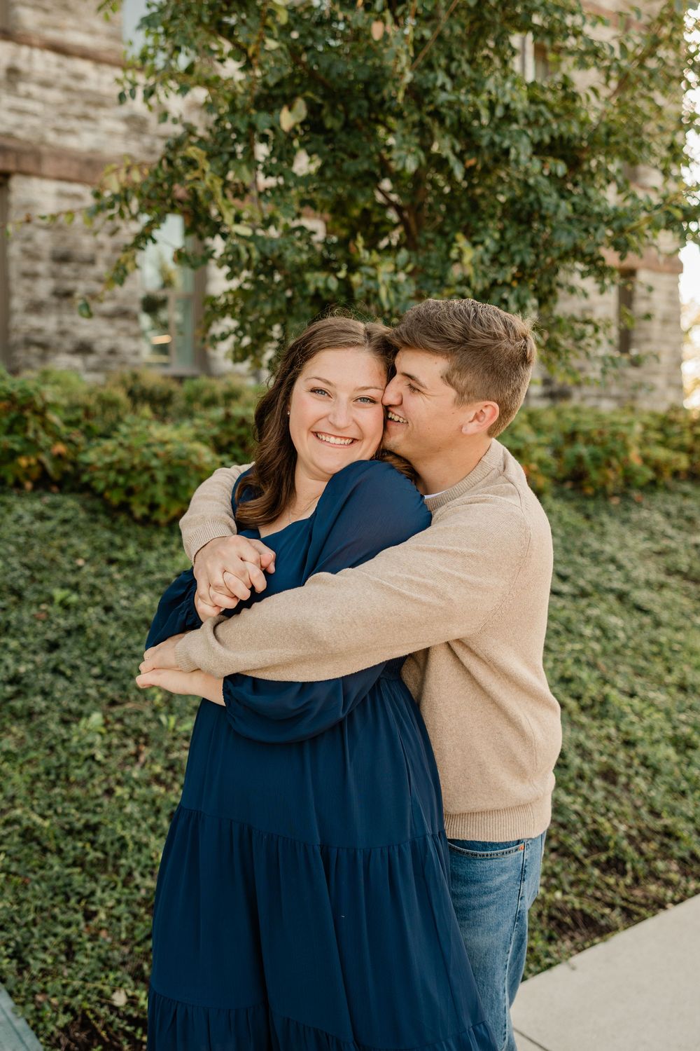 A couple embraces lovingly against a stone building backdrop with green foliage.