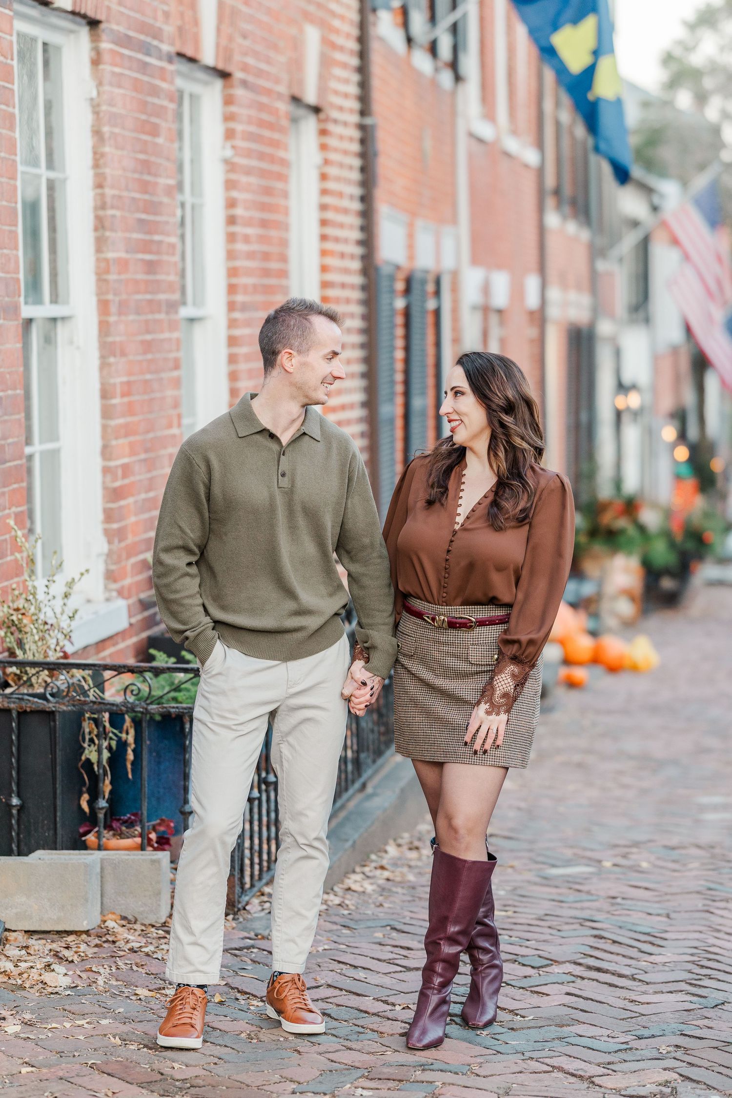 Couple walking together on a cobblestone street with fall decorations and brick buildings in the background.