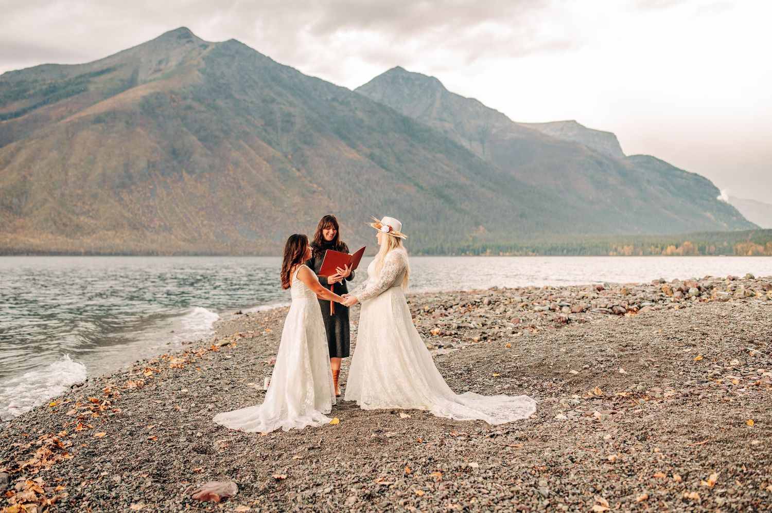 Three people in flowing dresses stand together on a rocky beach with dramatic mountain peaks in the background.