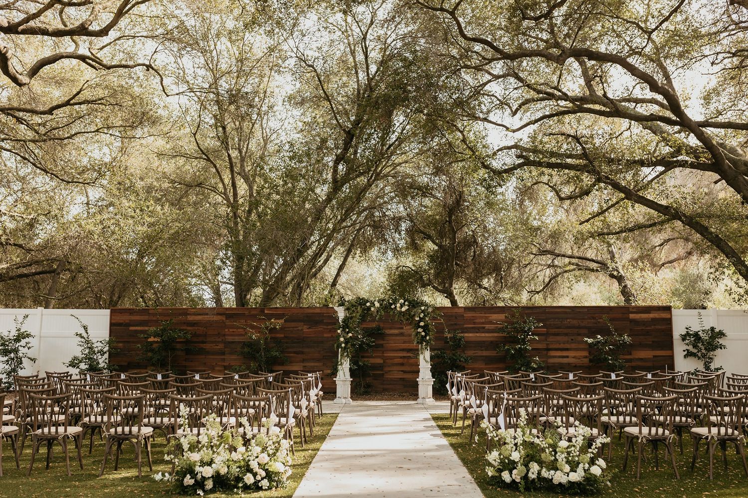 Rustic outdoor wedding ceremony setup with wooden benches and a white altar under tall trees in a garden setting at The Oaks at Duncan Lane.