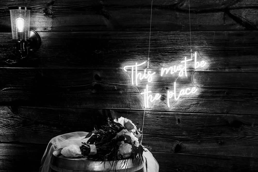 A neon sign reading 'Too much of a good thing' glows above a dark wooden wall in black and white photography.
