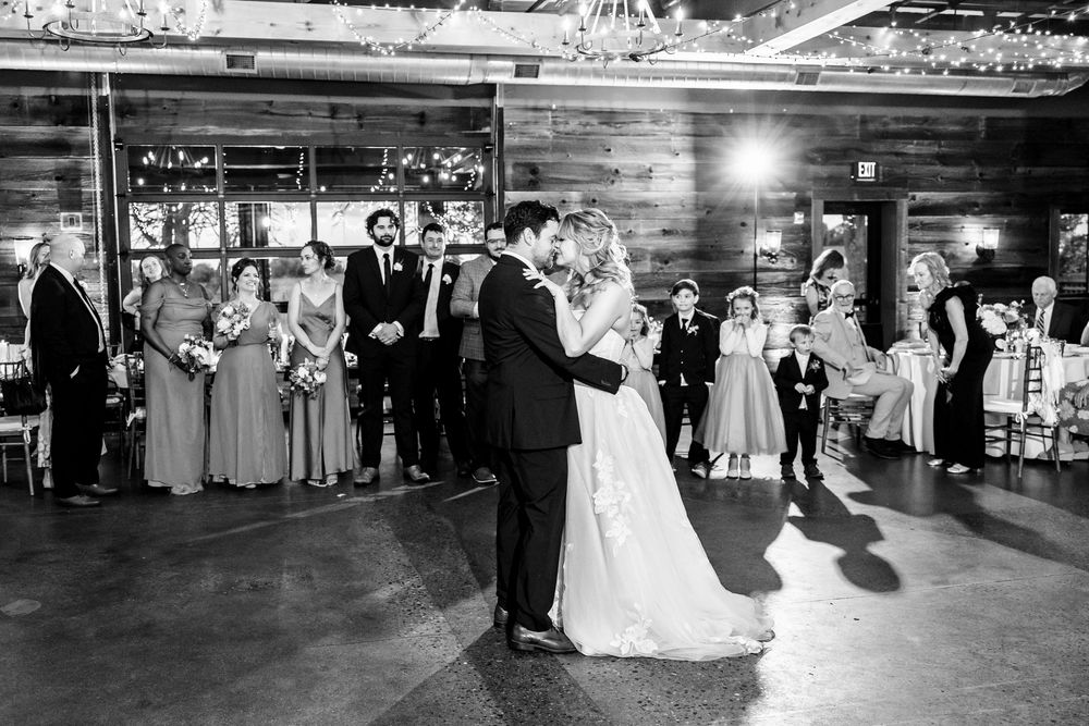 A black and white wedding reception photo showing a couple dancing while guests watch from the sides.