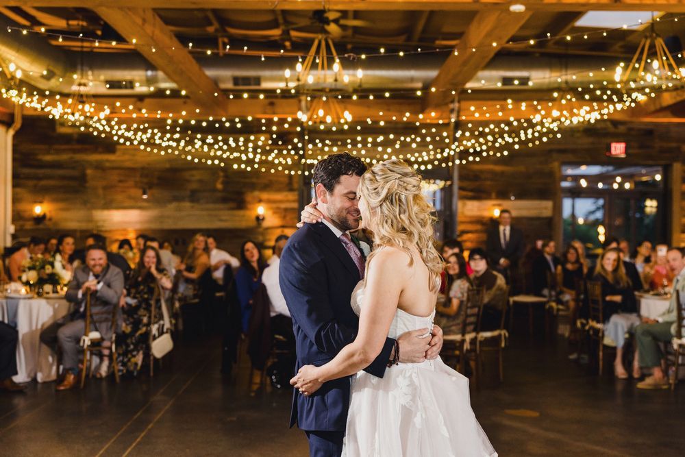 Newlyweds share an intimate dance moment under warm string lights at their rustic reception venue.
