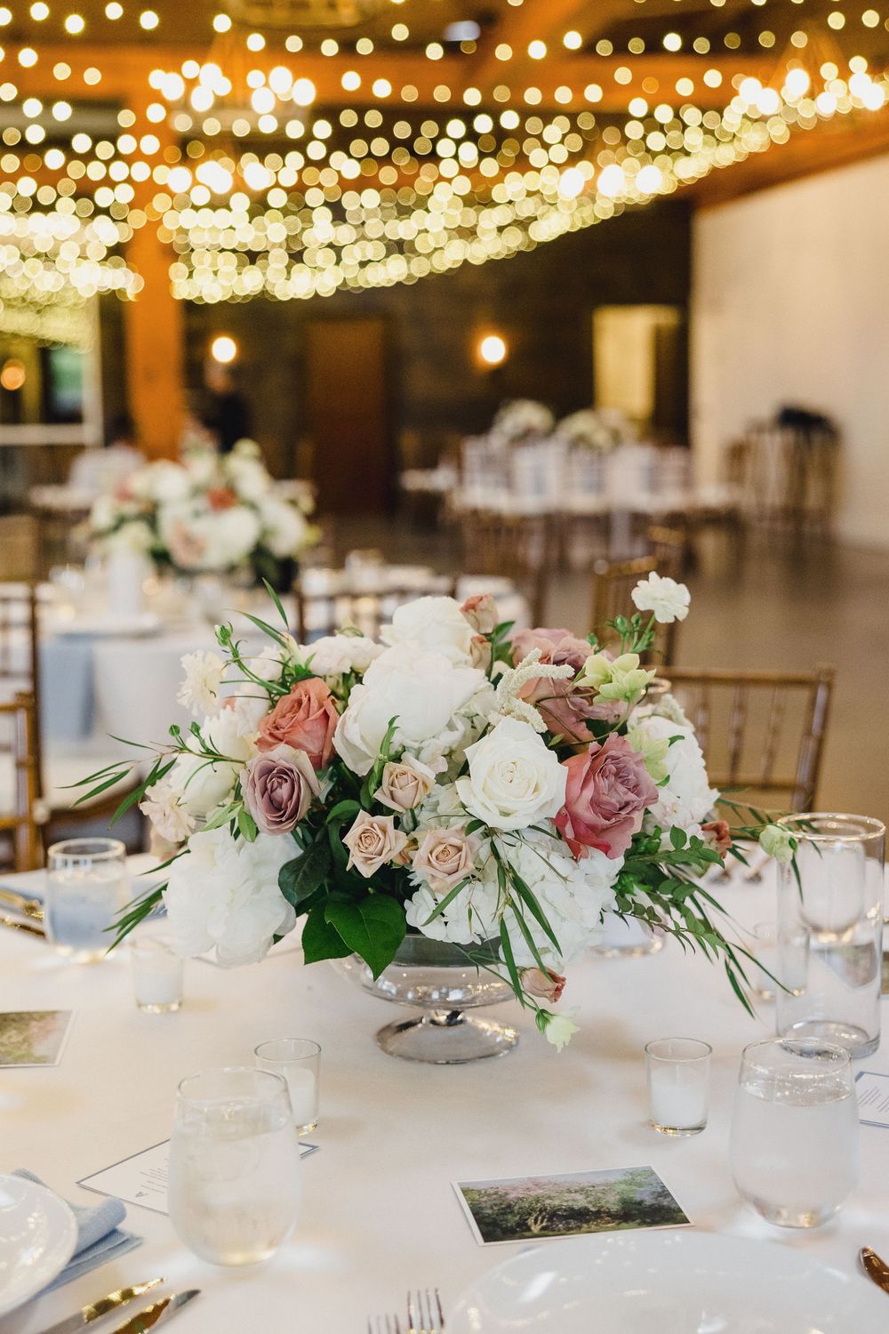 Elegant wedding reception table with white and pink floral centerpieces under warm string lights in a rustic venue.