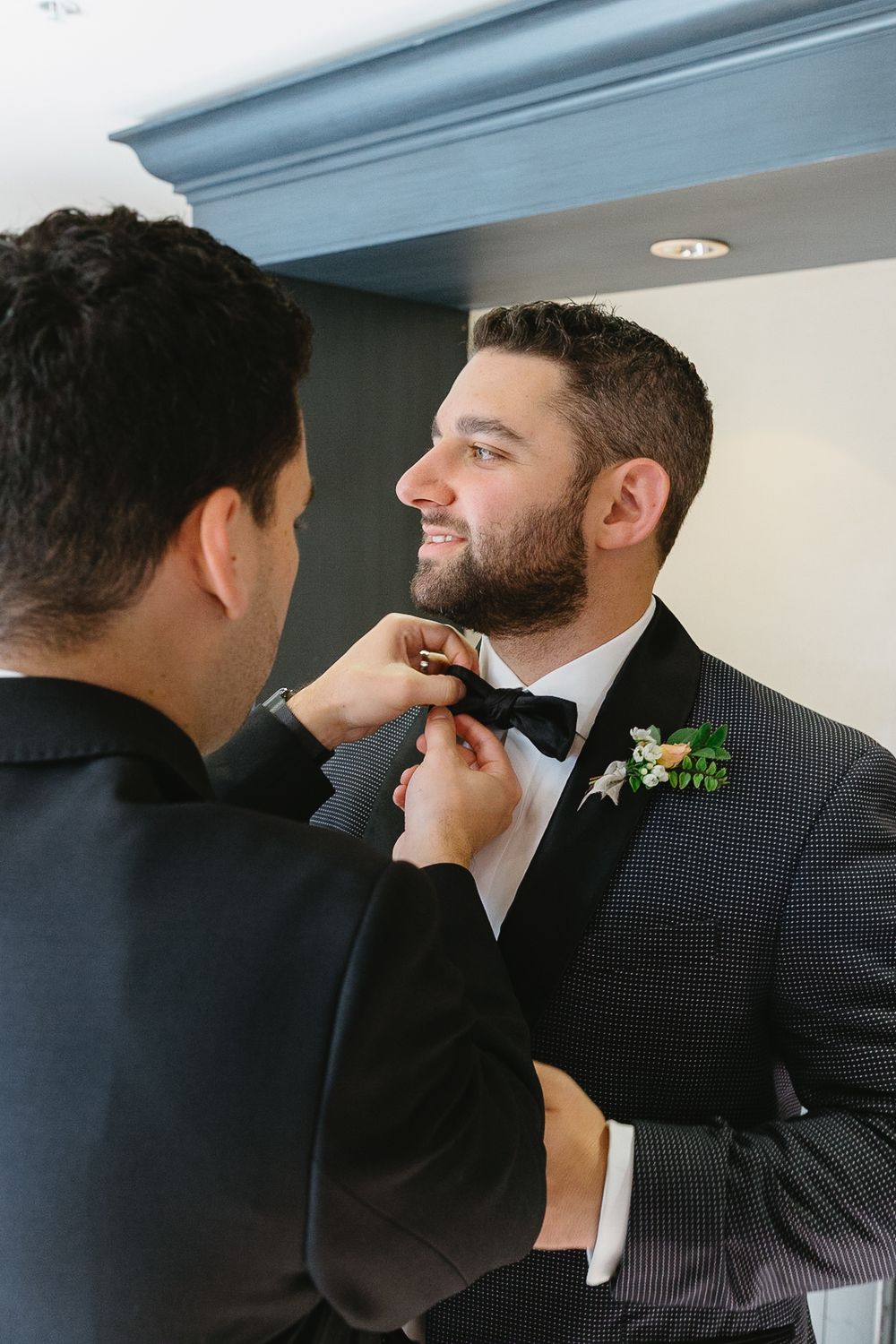 A groomsman helps adjust another man's tie and boutonniere before a formal event.