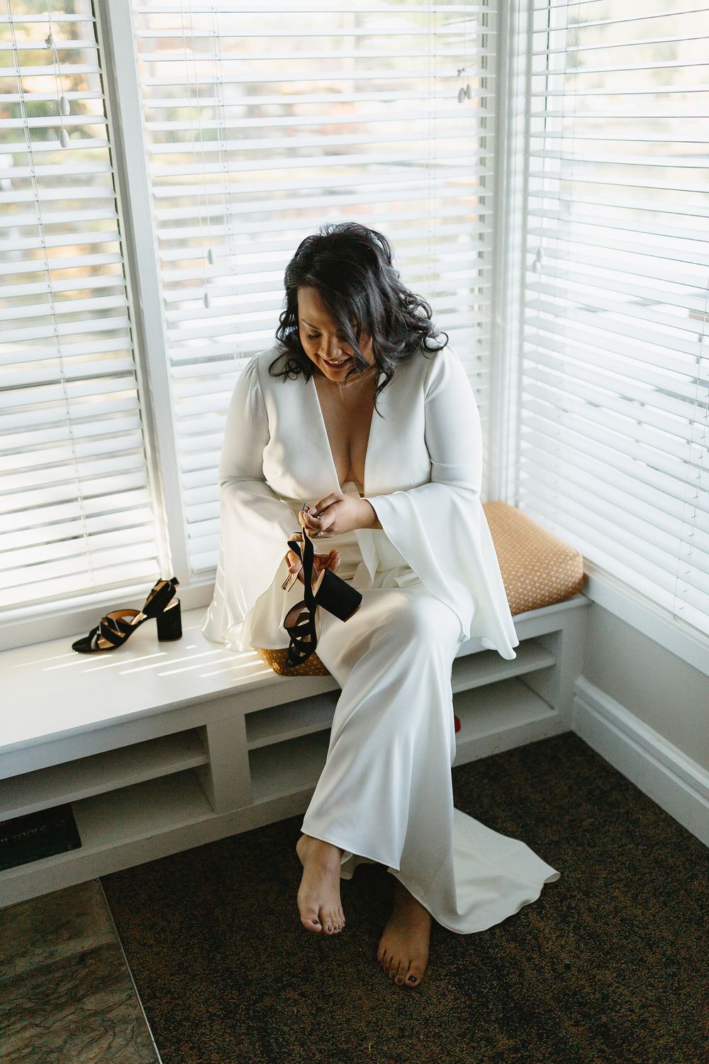A person in a white dress sits by a sunlit window with venetian blinds in a contemplative pose.