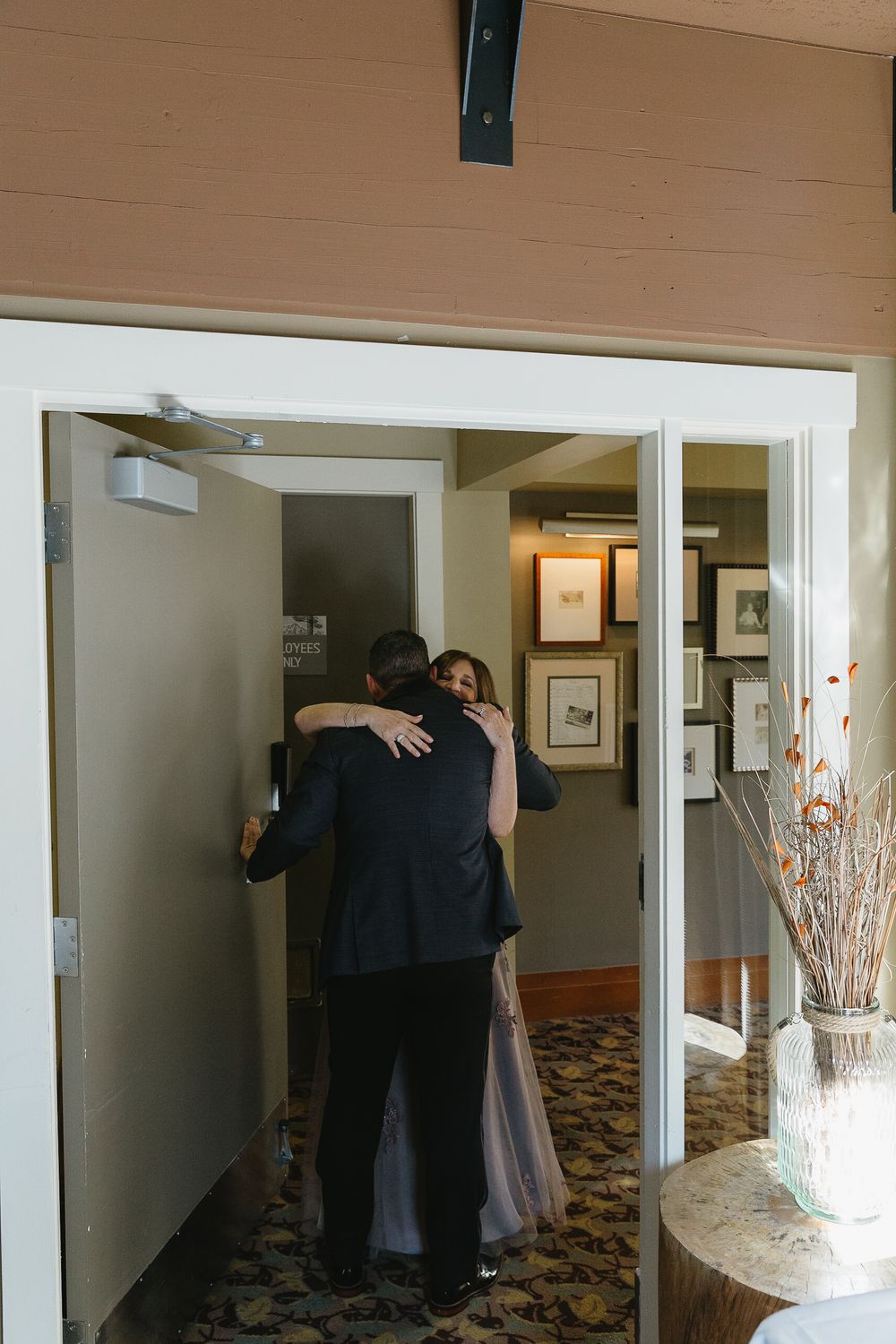 Two people embrace in a dimly lit hallway with framed artwork on the walls.