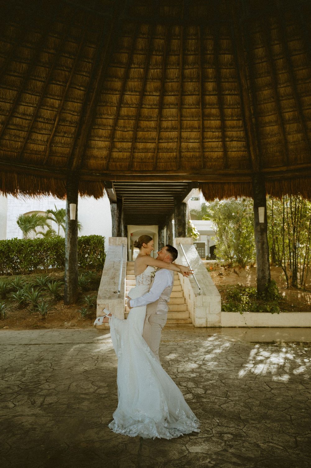 A couple shares a romantic dance under a thatched palapa roof at a tropical resort during golden hour.