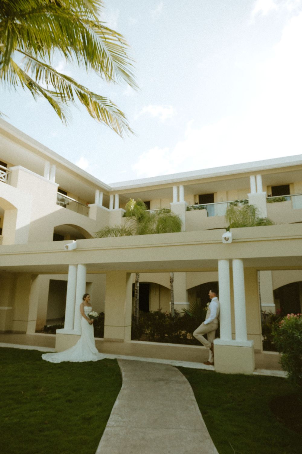 A couple in wedding attire poses at a luxury resort with white architecture and palm trees against a blue sky.