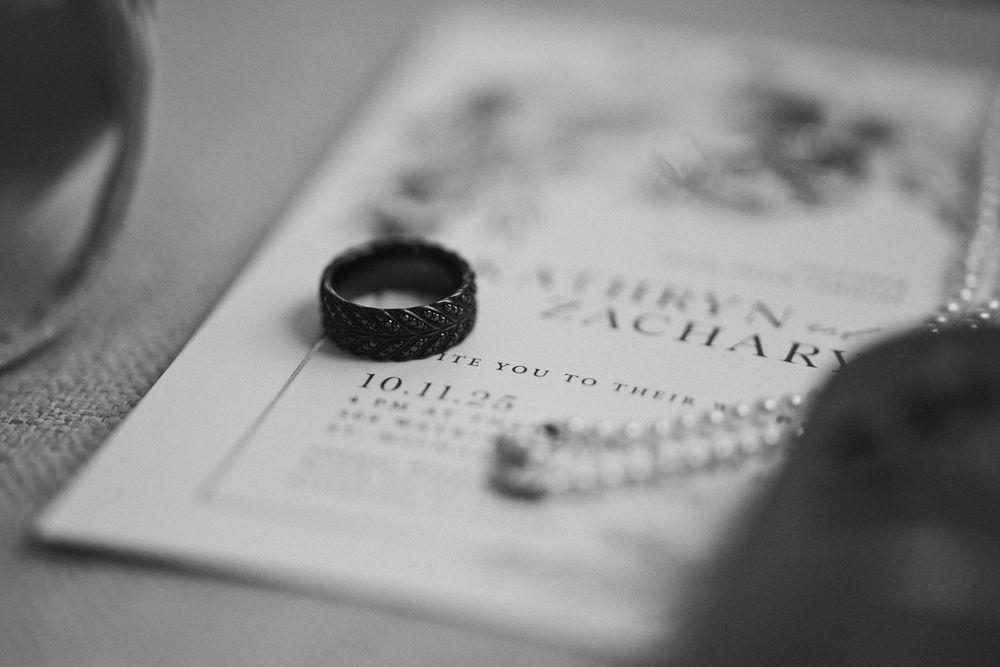 Black and white close-up of dark wedding band resting on wedding invitation dated 10.11.22.