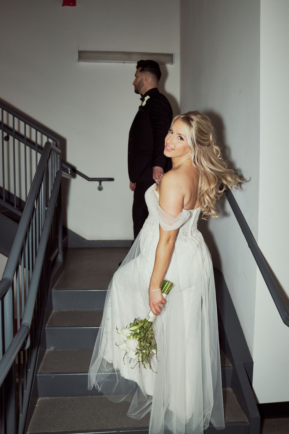 Smiling blonde bride in flowing white gown holds bouquet while ascending staircase with groom ahead of her.