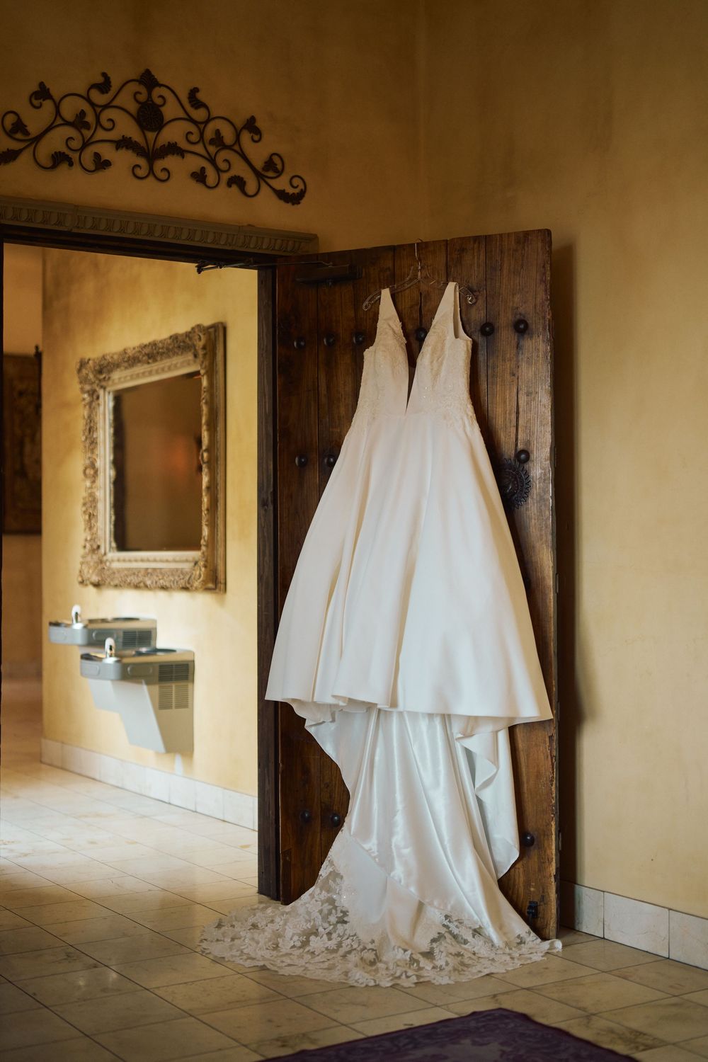 White wedding gown hanging on rustic wooden door in elegant venue with ornate mirror.
