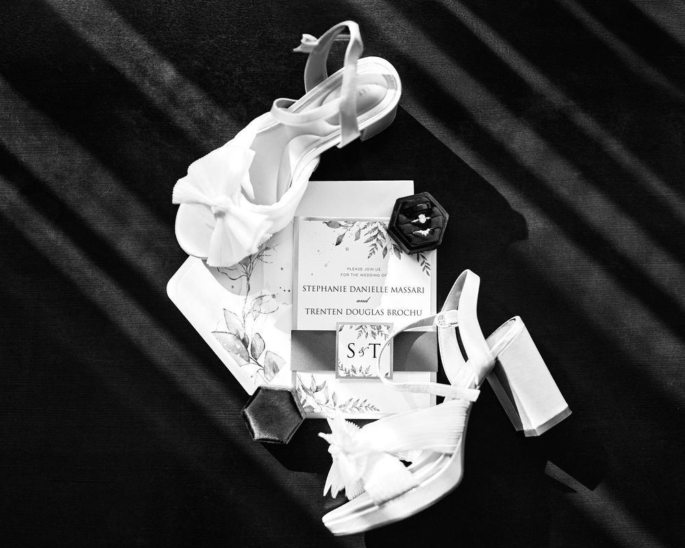 Black and white flat lay of white bridal heels, wedding invitation, and ring box.