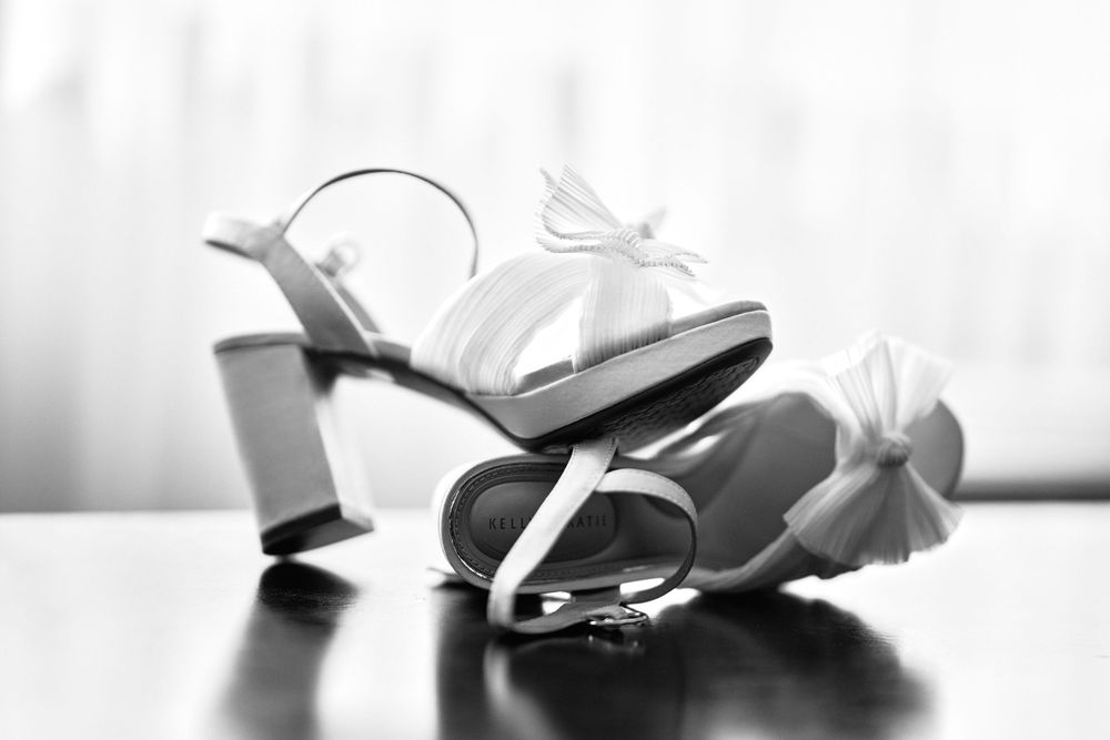 Black and white close-up of white block-heel bridal sandals with bow detail.