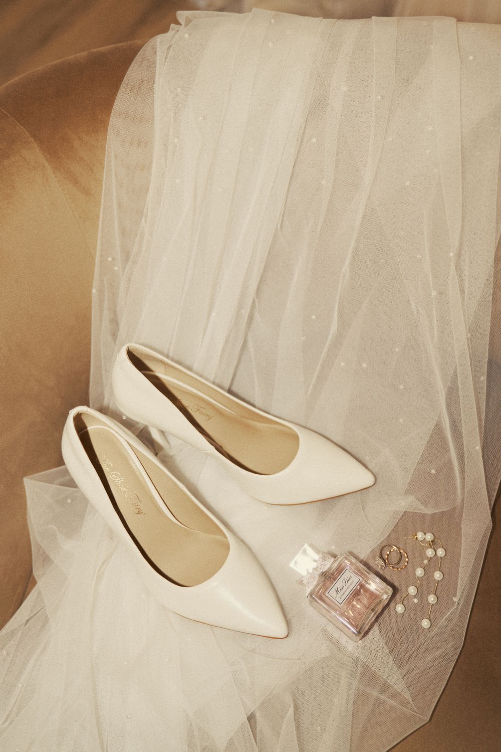 White pointed-toe bridal heels displayed on tulle fabric with perfume bottle and pearl earrings.