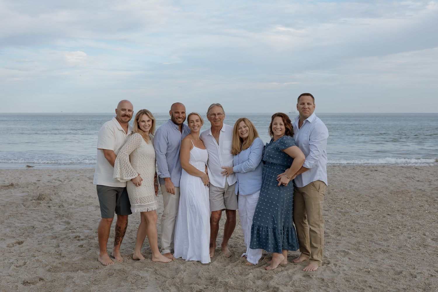 A group in white attire poses for photos at sunset on a beach with rolling waves and cloudy skies in the background.