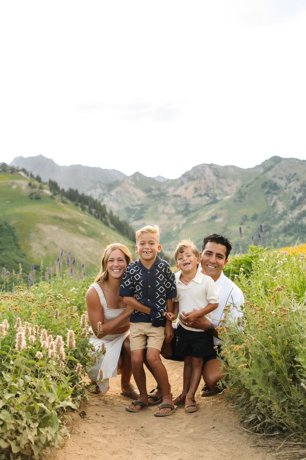 A family sits together on a mountain trail surrounded by wildflowers and scenic mountain peaks in the background for photos at Albion Basin.