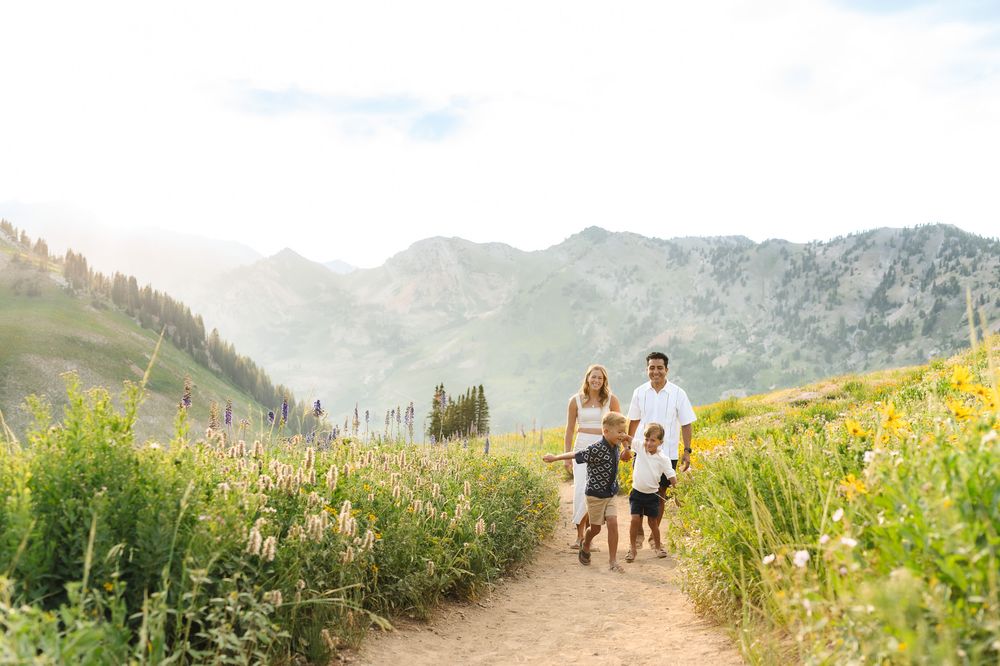 A couple and their dog hike along a scenic mountain trail surrounded by wildflowers and alpine peaks.