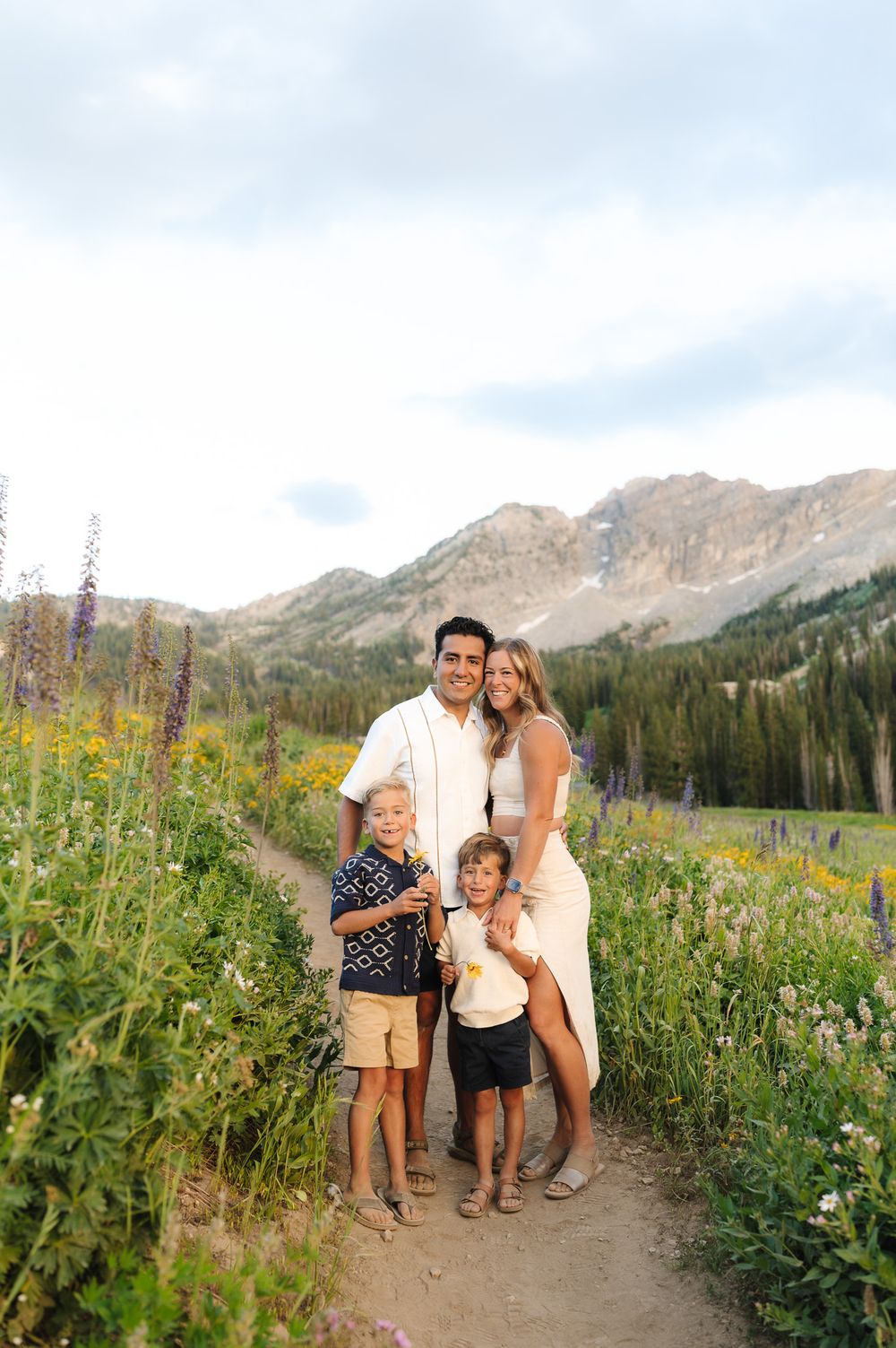 A family poses together on a hiking trail with wildflowers and mountain peaks in the scenic backdrop.