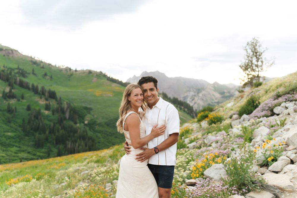 A couple embraces on a mountain trail surrounded by green alpine meadows and rocky peaks in the background.