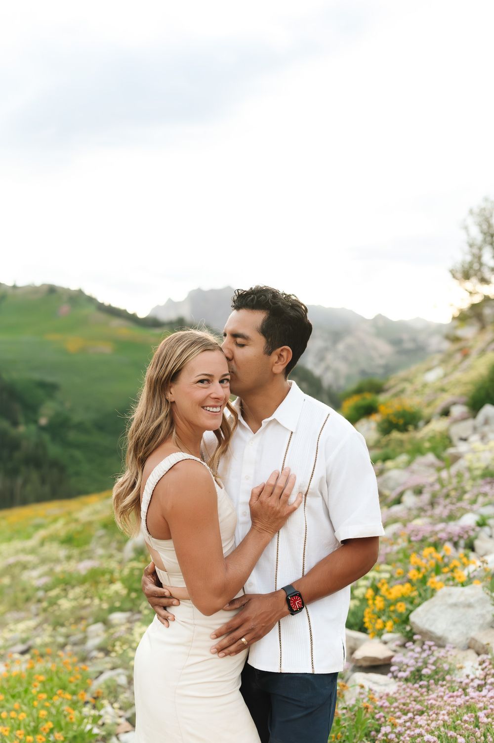 A romantic couple embraces and laughs together against a backdrop of mountain wildflowers and rolling green hills.