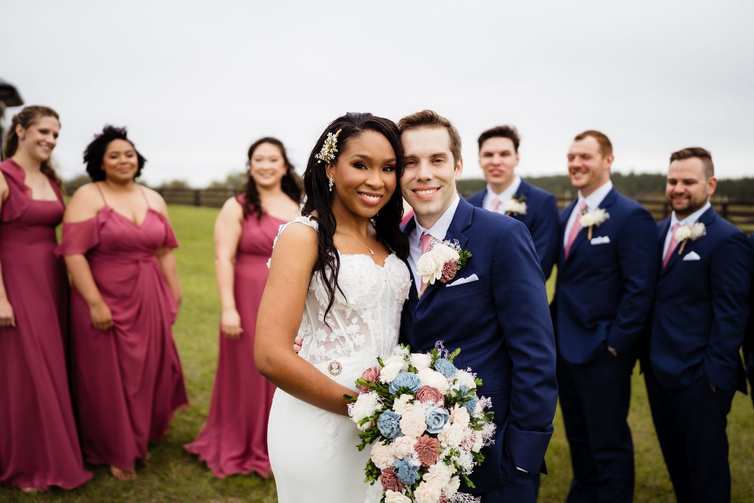 A wedding party poses outdoors wearing burgundy dresses and navy suits with pink and white floral bouquets.