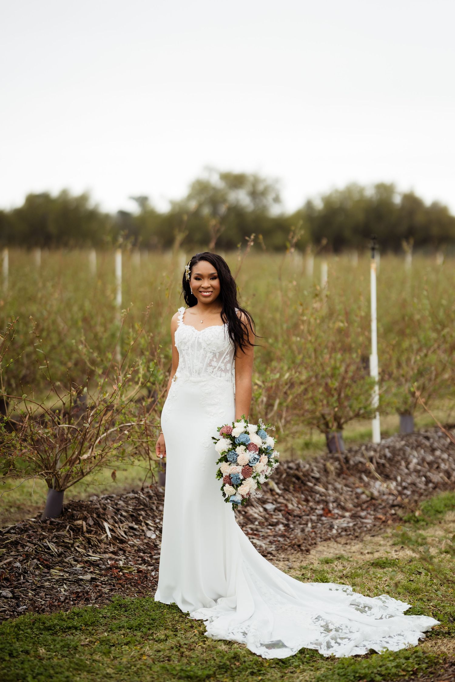 A bride in a flowing white lace gown poses elegantly in a vineyard during a rustic outdoor wedding photoshoot.