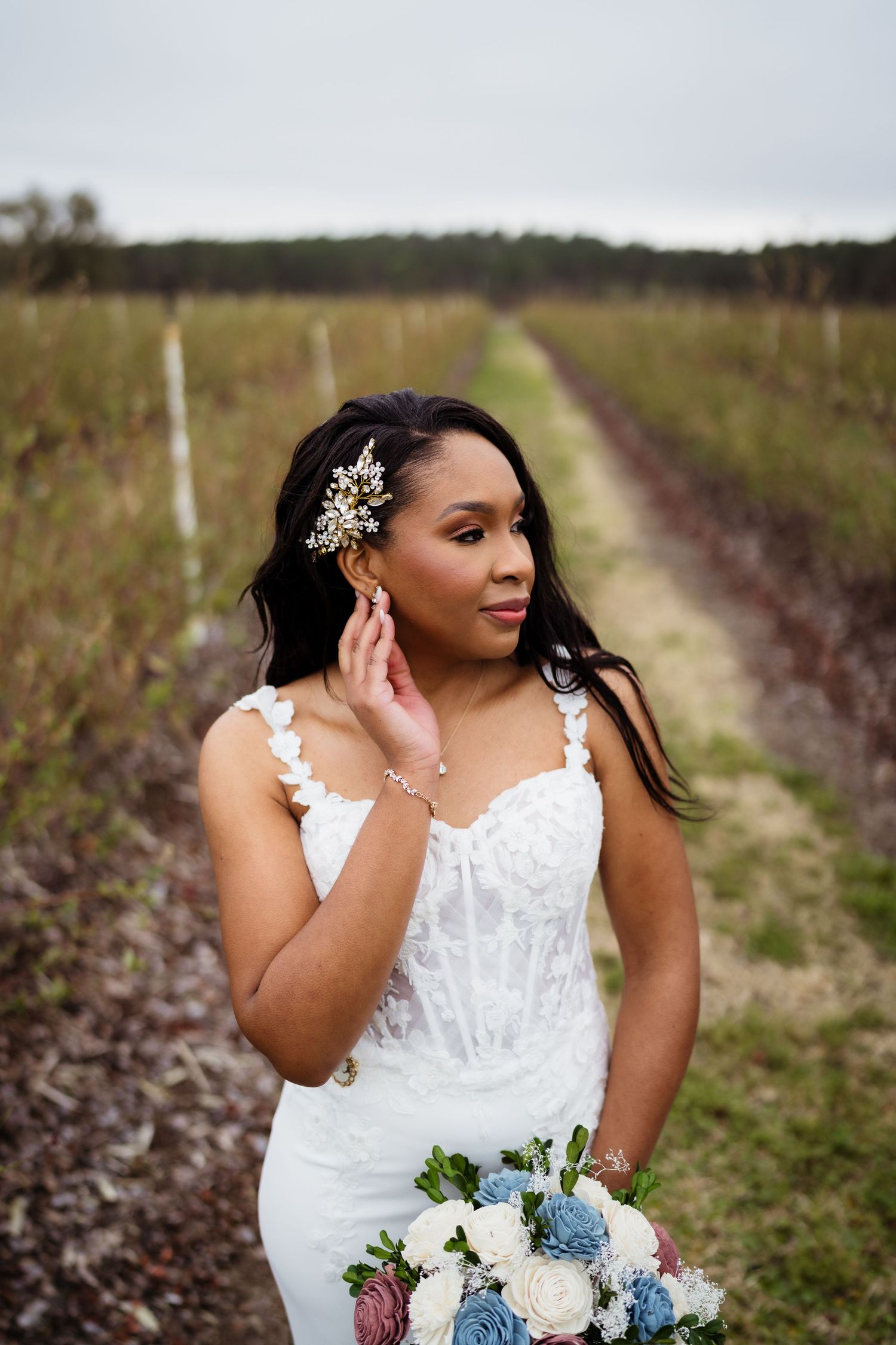 A bride in a white lace gown poses with a blue and white bouquet in a rustic outdoor setting.
