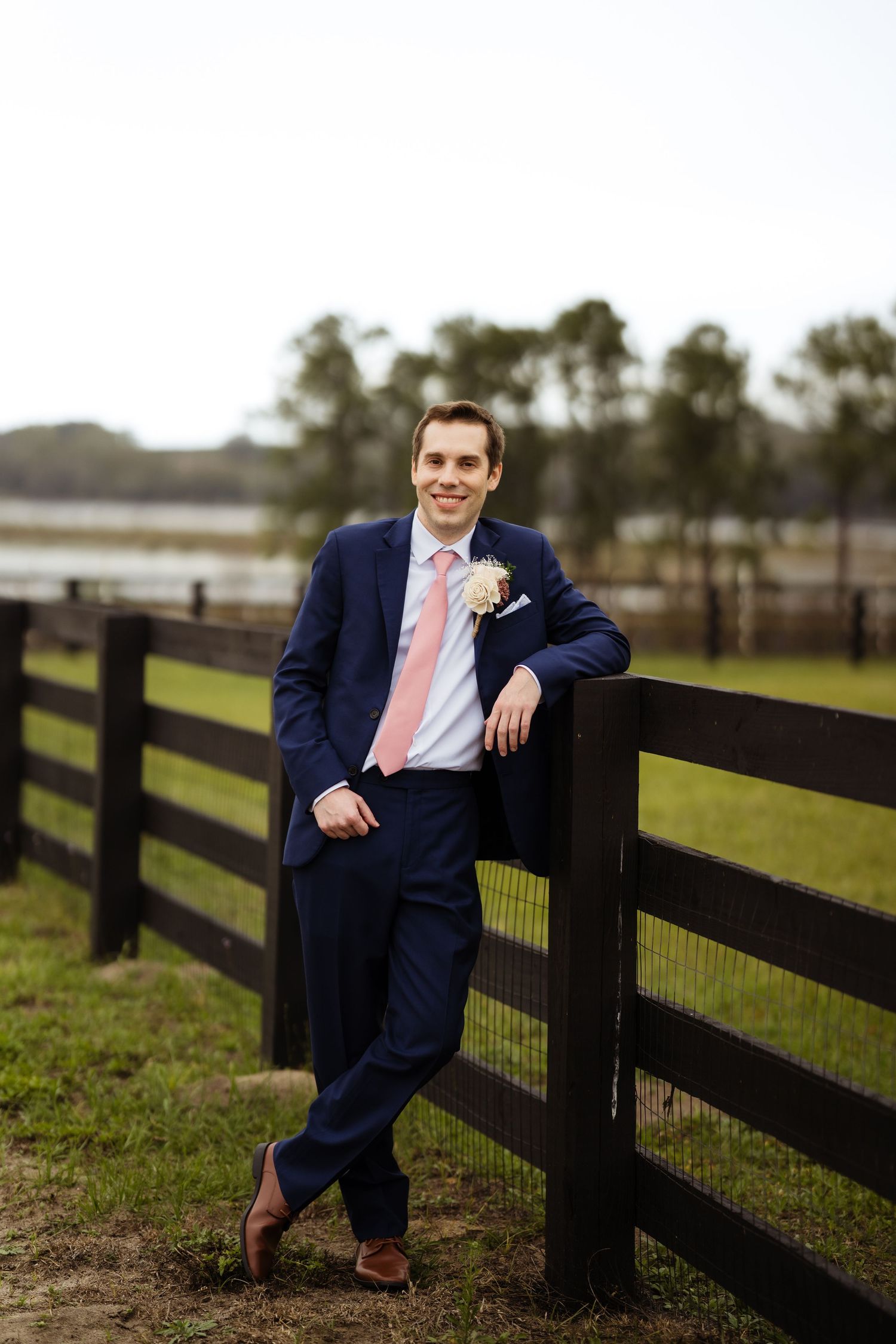 A groom in a navy suit and pink tie leans against a wooden fence at a countryside venue.