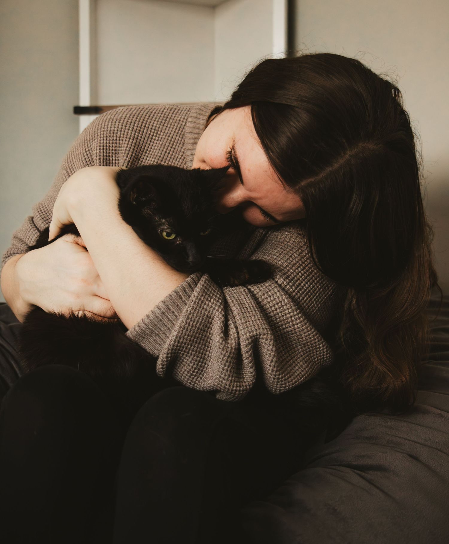 A person in a brown sweater lying down curled up on a bed in a dimly lit room.