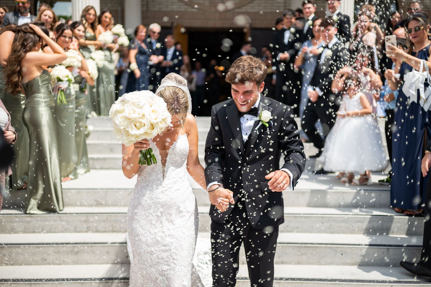 Wedding guests throw white confetti over a bride and groom as they exit down stone steps at their celebration.