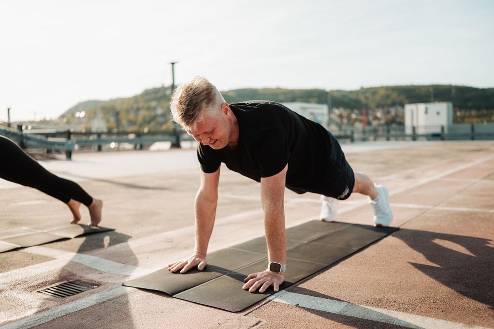 Person in black t-shirt doing plank exercise on outdoor workout mat during sunny day at beachfront location.